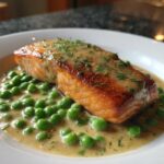A close-up of a perfectly cooked salmon fillet in a creamy dill sauce with green peas, served in a white bowl. Creamy Dill Salmon and Peas Skillet.