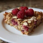 A close-up of a single serving slice of Healthy Raspberry Almond Baked Oatmeal on a white plate, topped with fresh raspberries and sliced almonds.