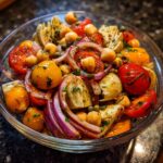 A close-up of a vibrant Artichoke Tomato Chickpea Salad in a glass bowl, featuring cherry tomatoes, chickpeas, artichoke hearts, and red onion.