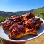 Close-up of juicy BBQ chicken thighs glazed with sauce, served on a white plate outdoors.