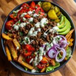 A close-up of a delicious Burger Bowl with baked fries, seasoned ground beef, tomatoes, avocado, red onion, and pickles, drizzled with sauce.