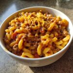 A close-up of a bowl of Classic American Goulash with Macaroni, topped with melted shredded cheese.