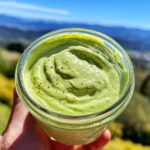 A close-up of a glass jar filled with vibrant, creamy avocado dressing, sprinkled with pepper, held outdoors.