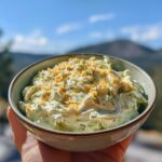 A bowl of creamy Dill Pickle Dip topped with fresh dill and crumbled cheese, held by a hand.