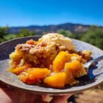 A close-up of a serving of Easy Peach Cobbler with Fresh Peaches topped with vanilla ice cream, held on a plate with a fork.
