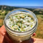 A hand holds a jar of creamy Garlic Yogurt Sauce for Everything, with fresh herbs and garlic visible.
