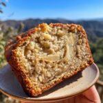 A close-up of a slice of moist Greek yogurt banana bread with visible banana pieces, held on a small plate.