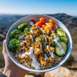 A hand holds a bowl of a healthy chicken bowl meal prep recipe with brown rice, chicken, broccoli, tomatoes, and cucumber, drizzled with sauce.