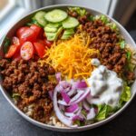 A vibrant High Protein Burger Bowl filled with seasoned ground beef, rice, lettuce, tomatoes, cucumber, avocado, cheese, and red onion, topped with a dollop of sour cream.