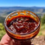 Close-up of a jar filled with rich, dark Homemade BBQ Sauce Easy Version, with a scenic mountain landscape in the background.