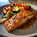 A glistening fillet of Honey Garlic Salmon served with a colorful medley of roasted vegetables including broccoli, bell peppers, zucchini, and carrots.