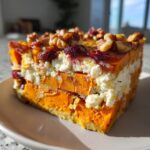 A slice of Layered Sweet Potato and Squash Vegetable Bake with layers of orange vegetables, white cheese, and topped with walnuts and cranberries.