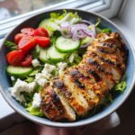 A vibrant bowl of Lemon Herb Grilled Chicken Salad with sliced grilled chicken, cherry tomatoes, cucumber, red onion, and feta cheese.