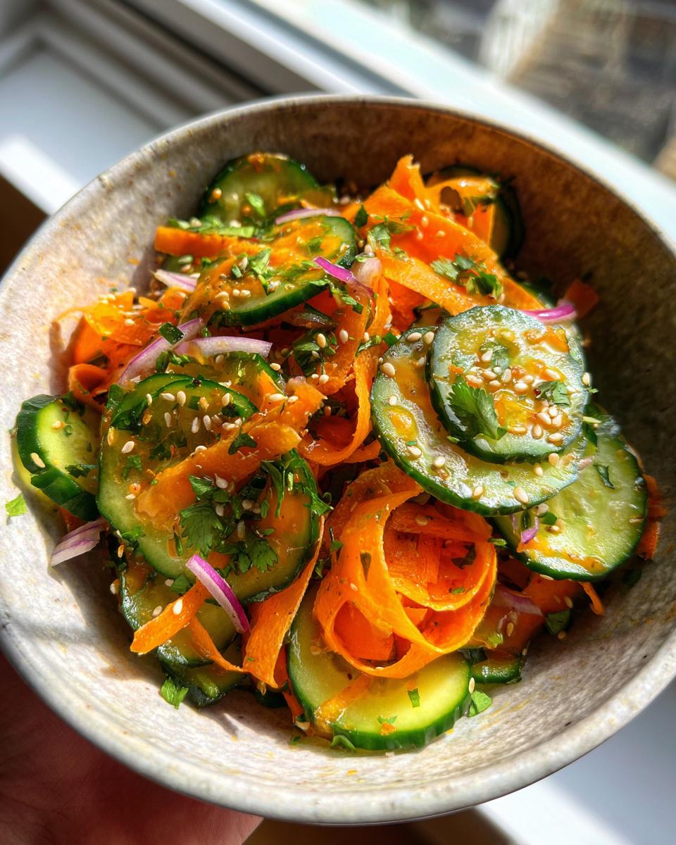 A close-up of a vibrant Low Calorie Cucumber Carrot Crunch Salad in a rustic bowl, featuring thinly sliced cucumbers, ribbons of carrots, red onion, and sesame seeds.