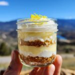 A hand holding a mason jar filled with a layered no bake lemon dessert, topped with whipped cream and lemon zest.