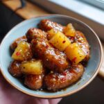 A close-up of Pineapple Brown Sugar Beef Smokies Bites in a bowl, glazed and topped with pineapple chunks and red pepper flakes.