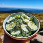 A bowl of refreshing cucumber yogurt salad with fresh dill, held by a hand, with a scenic mountain view in the background.