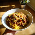 A hand holding a white bowl filled with Spaghetti Squash Burrito Bowls topped with seasoned ground beef, beans, melted cheese, and green onions.
