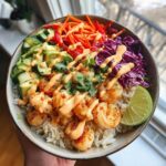 A vibrant bowl of Spicy Shrimp Rice Bowls topped with creamy chili sauce, avocado, cucumber, carrots, and red cabbage.