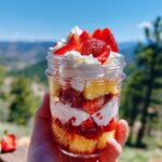 A hand holds a mason jar filled with layers of cake, strawberries, and whipped cream, representing an easy Strawberry Shortcake Cups dessert.