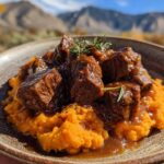 A close-up of tender Apple Cider Beef served over a bed of creamy mashed sweet potatoes, garnished with fresh herbs.