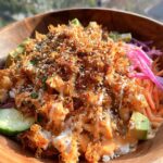 A close-up of a bang bang chicken crispy rice salad bowl with avocado, carrots, and pickled onions, drizzled with sauce.