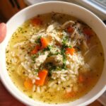 A close-up of a bowl of delicious chicken and rice soup, featuring shredded chicken, white rice, and diced carrots, garnished with fresh parsley.