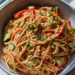 A close-up bowl of cold peanut noodles tossed with shredded carrots, sliced cucumbers, red bell peppers, and chopped peanuts.