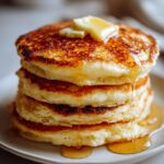 A tall stack of golden-brown cottage cheese pancakes topped with melting butter and drizzled with syrup.