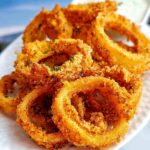 A pile of golden brown, crispy keto onion rings coated in breadcrumbs and herbs, served on a white plate.