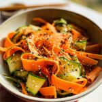 Close-up of a bowl filled with ribbons of cucumber and carrot, tossed in a sesame dressing and sprinkled with sesame seeds.