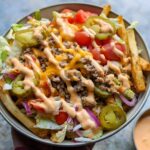 A bowl filled with an Easy Smash Burger Bowl Recipe, featuring seasoned fries, ground beef, cheese, lettuce, tomatoes, onions, jalapeños, and a creamy sauce.
