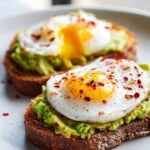 Two slices of perfect egg avocado toast topped with a fried egg and sprinkled with chili flakes.