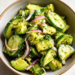 A close-up shot of a Fresh Cucumber Avocado Salad in a bowl, featuring sliced cucumbers, creamy avocado, red onion, and herbs.