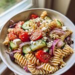 Close-up of a bowl of Greek pasta salad with feta, featuring rotini pasta, cherry tomatoes, cucumber, red onion, and olives.