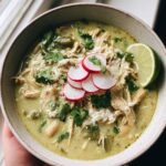A close-up of a bowl of green enchilada chicken soup, garnished with radishes, cilantro, cheese, and a lime wedge.