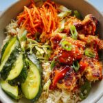 A close-up of a Healthy Bang Bang Chicken Bowl featuring crispy chicken coated in creamy spicy sauce, rice, shredded carrots, and sliced cucumbers.