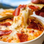 A hand dips a chip into a bubbly High Protein Cottage Cheese Pizza Bowl topped with melted cheese and pepperoni.