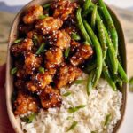 A bowl of High Protein Sesame Chicken served with steamed rice and fresh green beans, garnished with sesame seeds and scallions.