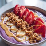 Close-up of a vibrant purple high protein smoothie bowl topped with sliced strawberries, banana, granola, and a drizzle of peanut butter.