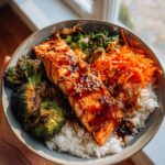 A close-up of a honey garlic salmon bowl with rice, roasted broccoli, and shredded carrots, drizzled with sauce and sesame seeds.