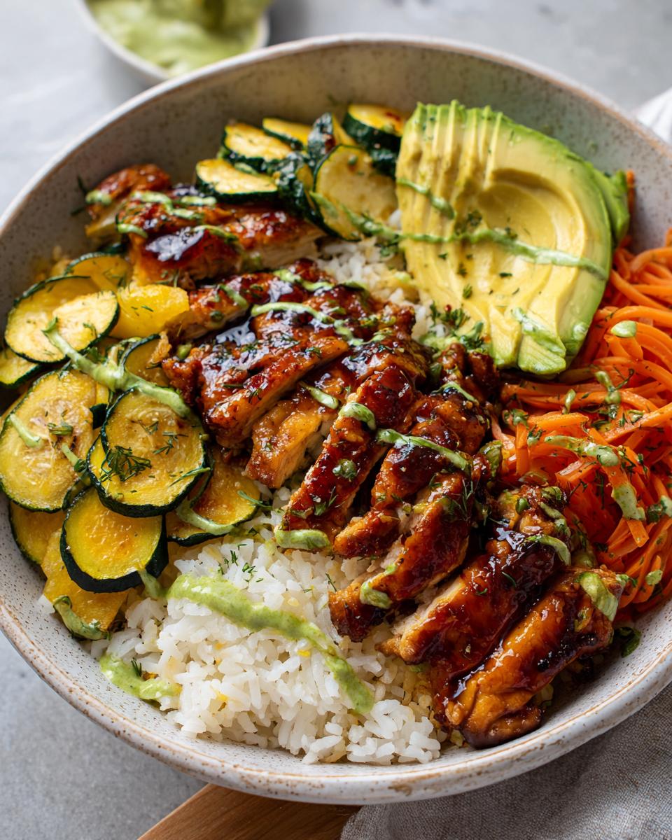 A close-up of a hot honey chicken rice bowl topped with sliced avocado, zucchini, and shredded carrots, drizzled with green sauce.
