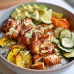 A delicious hot honey chicken rice bowl topped with sliced avocado, lime, shredded carrots, and zucchini.