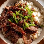 A close-up of a bowl of fluffy white rice topped with tender slices of Mongolian beef in a glossy sauce, garnished with fresh green onions.