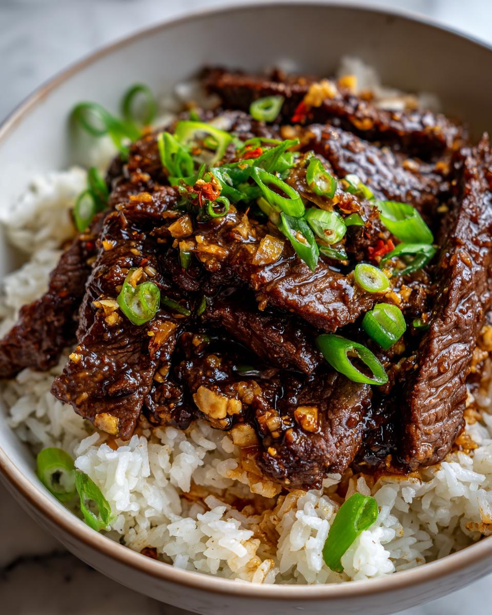 Close-up of a bowl of fluffy white rice topped with tender slices of savory Mongolian beef and garnished with chopped green onions and chili flakes.