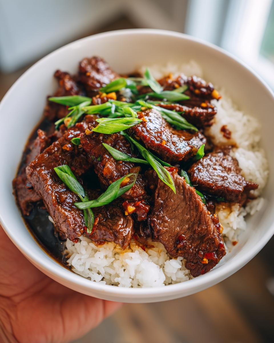 A close-up of a white bowl filled with fluffy white rice topped with tender slices of Mongolian beef and garnished with chopped green onions.