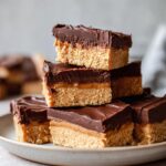 A stack of no bake chocolate peanut butter bars featuring a crunchy base, creamy peanut butter layer, and rich chocolate topping.