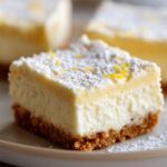 Close-up of a no bake lemon cheesecake bar with a graham cracker crust, dusted with powdered sugar and lemon zest.
