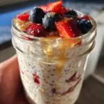 A close-up of protein overnight oats in a jar, topped with fresh strawberries, blueberries, chia seeds, and a drizzle of honey.