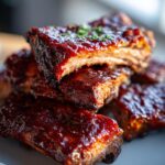 A close-up stack of tender slow cooker BBQ ribs coated in a rich, glossy sauce, garnished with fresh herbs.